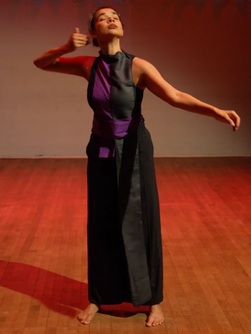 Varsha in a purple and black costume, eyes closed, one arm raised behind her head and the other reaching out, on a wooden floor in red light