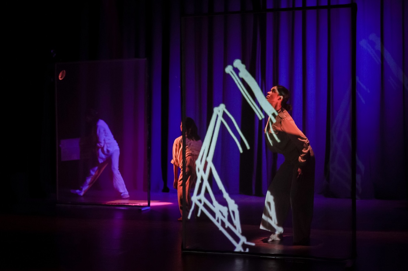 Performers on a dark stage between two framed screens, a white line drawing projected across one of them, blue and violet light